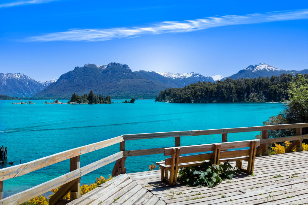 Wooden viewing deck on Isla Victoria overlooking the vivid turquoise waters of Lago Nahuel Huapi, with forested islands and snow-capped Andes peaks in Nahuel Huapi National Park, Bariloche, Argentina.