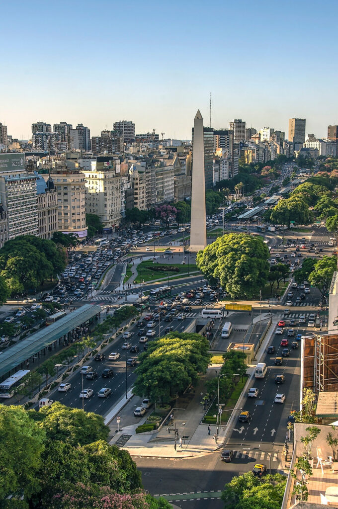Aerial view looking north along Avenida 9 de Julio in Buenos Aires, Argentina, with the 67-metre Obelisco de Buenos Aires monument rising from Plaza de la República amid heavy city traffic.