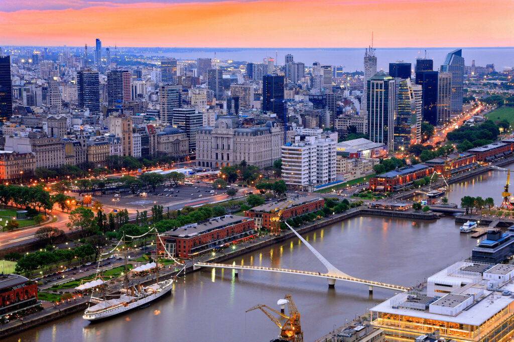 Aerial view of Puerto Madero waterfront at sunset in Buenos Aires, Argentina, with the illuminated Puente de la Mujer footbridge, the ARA Sarmiento museum ship and the Río de la Plata glowing under an orange sky.