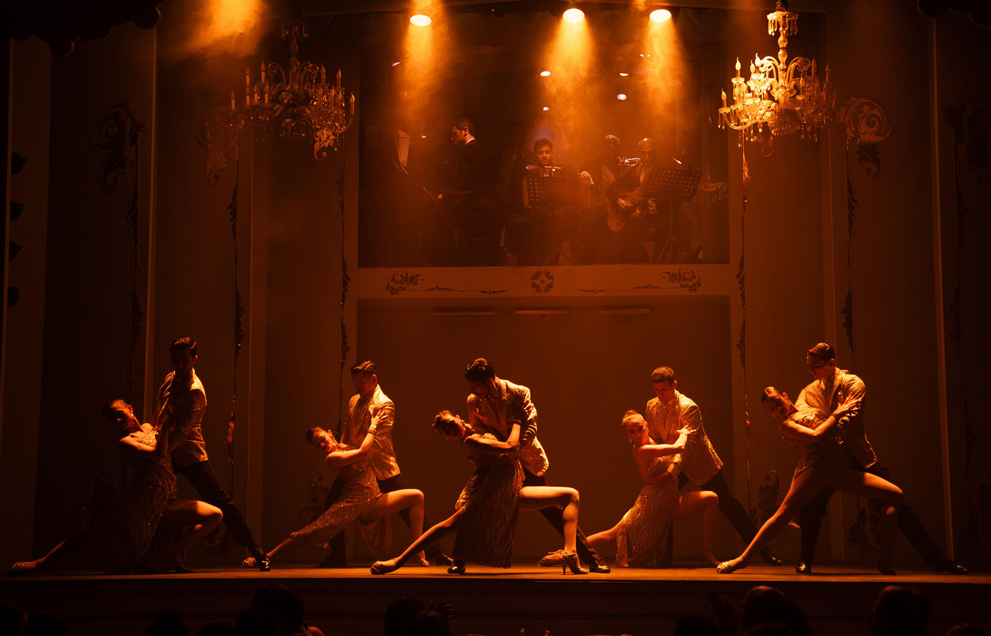 Professional tango show at Café de los Angelitos in Buenos Aires, Argentina, with four couples performing dramatic close-embrace corte and volcada figures on stage under warm amber theatrical lighting, crystal chandeliers and a live orquesta típica in the mezzanine above.