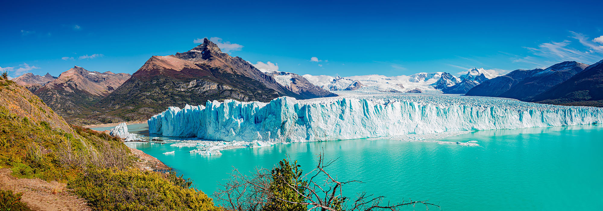 Panoramic view of Perito Moreno Glacier's vast blue-white ice wall calving into the turquoise waters of Lago Argentino in Los Glaciares National Park, Argentine Patagonia.