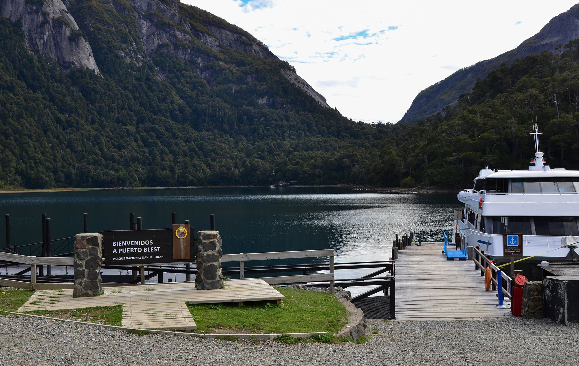 Puerto Blest welcome sign and wooden boat dock in Nahuel Huapi National Park, Argentina, with a lake cruise catamaran moored alongside and a steep fjord-like valley of Valdivian temperate rainforest rising on both sides of the glacial lake arm.