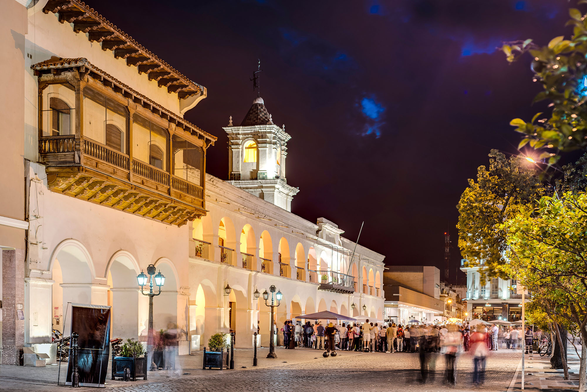 Cabildo of Salta illuminated at night on Plaza 9 de Julio in Salta, Argentina, with colonnaded white arches, carved wooden balconies and crowds gathering on the cobblestone square.