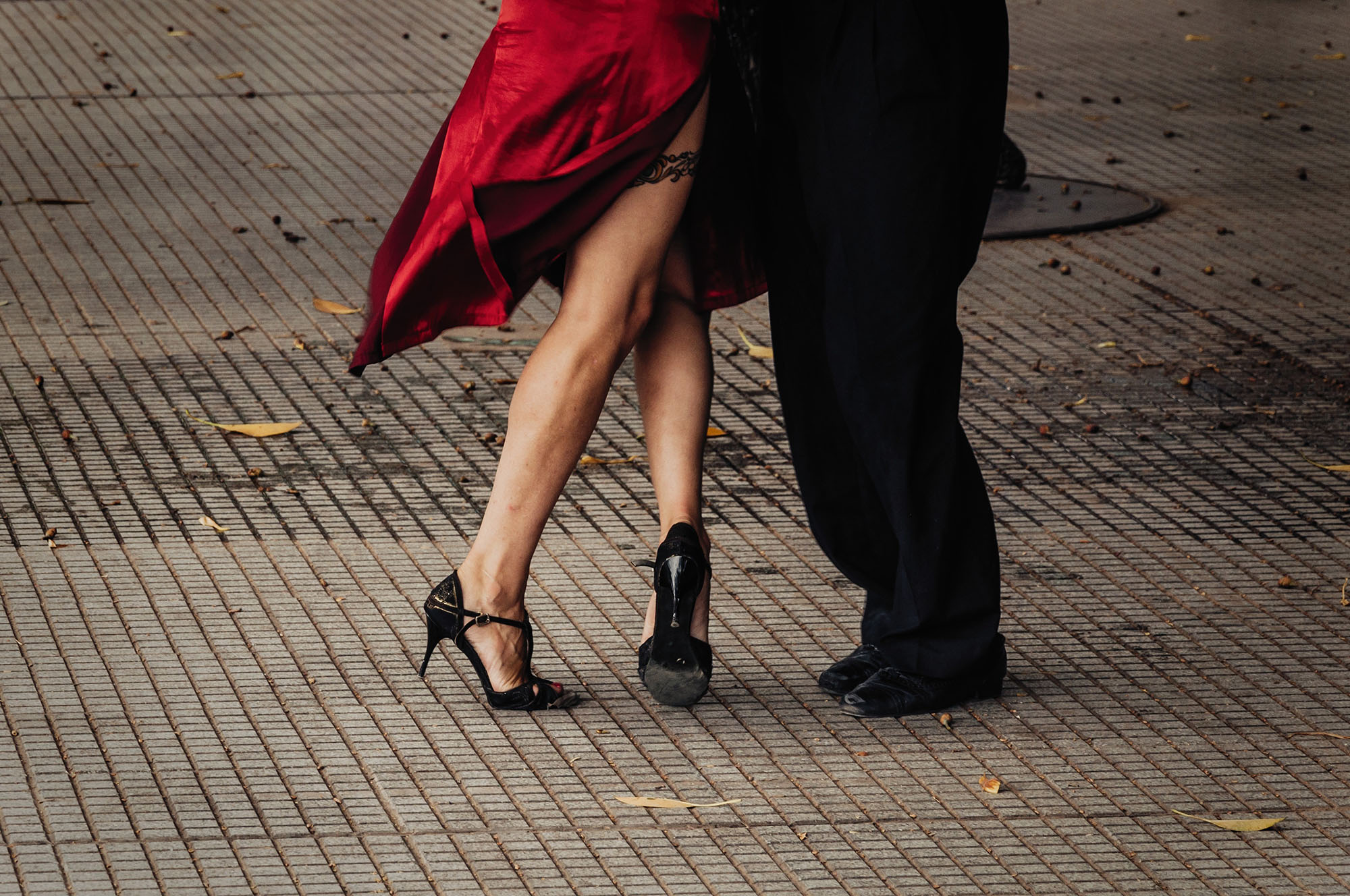 Close-up of Argentine tango dancers' feet and legs mid-step on a Buenos Aires street pavement, with a woman in a flowing red silk dress and black stiletto tango heels performing a gancho with her male partner.