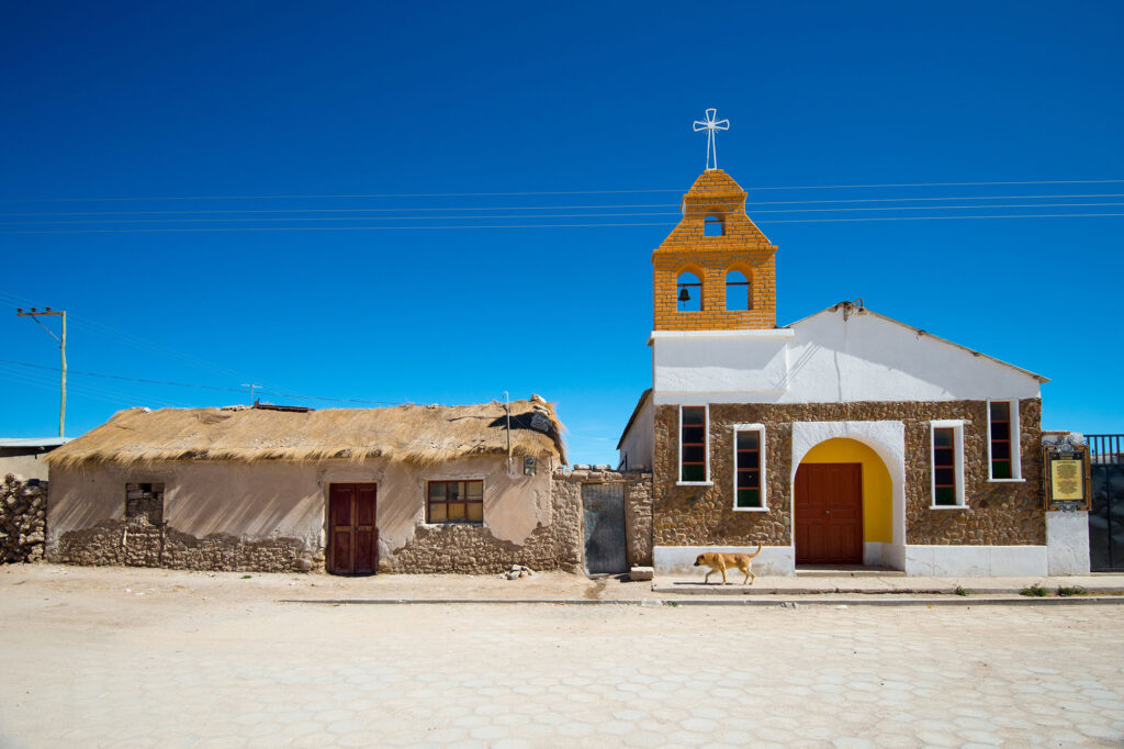 The whitewashed stone Catholic church with a yellow ochre brick bell tower in the remote altiplano village of Colchani, Bolivia, beside a traditional ichu grass-thatched adobe dwelling on a salt-flat cobblestone street, near the entrance to the Salar de Uyuni.