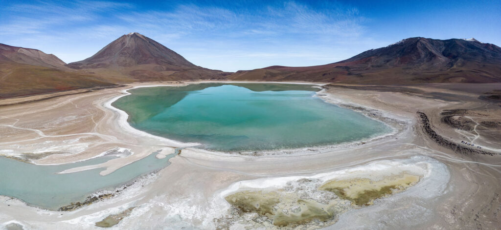 Aerial panoramic view of Laguna Verde — Green Lagoon — on the Bolivian altiplano near the Chilean border, with its vivid turquoise mineral-rich water, white salt and mineral shoreline and the perfectly conical Volcán Licancabur rising to the left above the high-altitude desert basin.