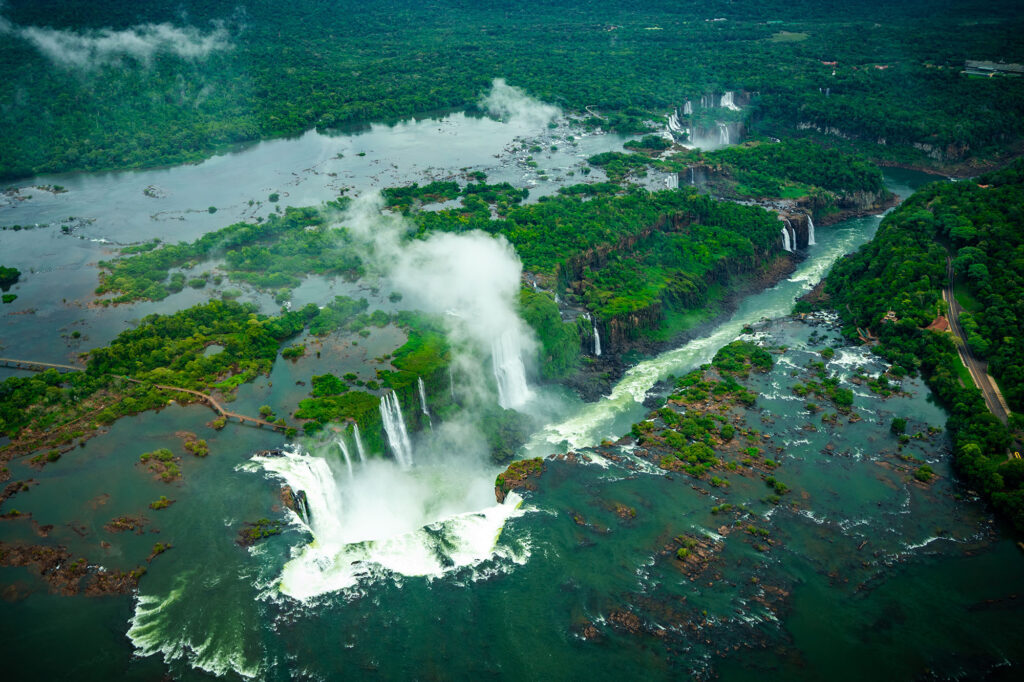 Helicopter aerial view of the complete Iguaçu Falls system from the Brazilian side, showing the Garganta do Diabo at peak flood season surrounded by submerged Atlantic Forest islands, with a towering mist plume rising above the Iguaçu River gorge.