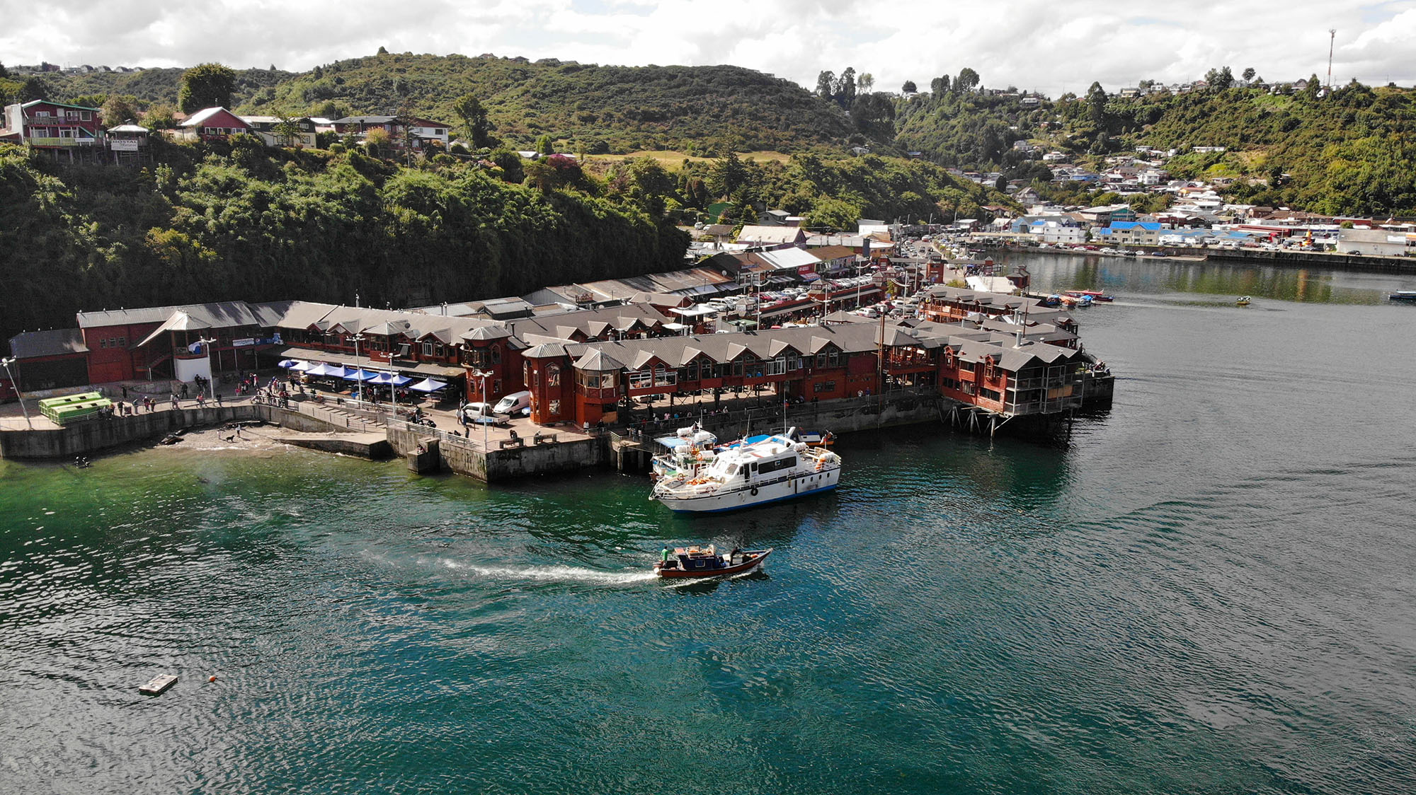 Aerial view of Mercado Angelmó fish market on the waterfront of Puerto Montt, Los Lagos Region, Chile, with fishing boats moored alongside the red-roofed timber market buildings extending over the turquoise water on stilts.