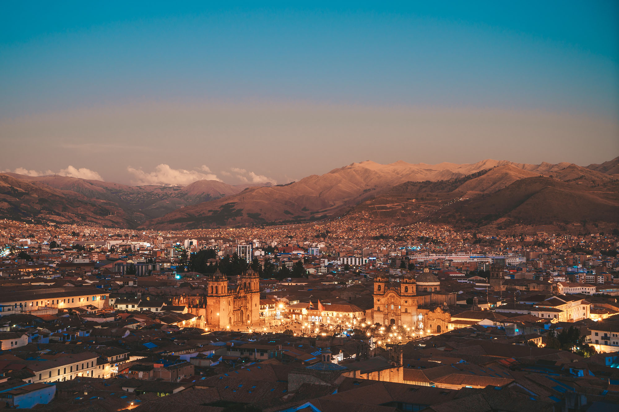 Aerial panoramic view of Cusco city at dusk, with the illuminated Plaza de Armas and Cusco Cathedral glowing at the centre, surrounded by terracotta rooftops and the Andean mountains at twilight, Peru.