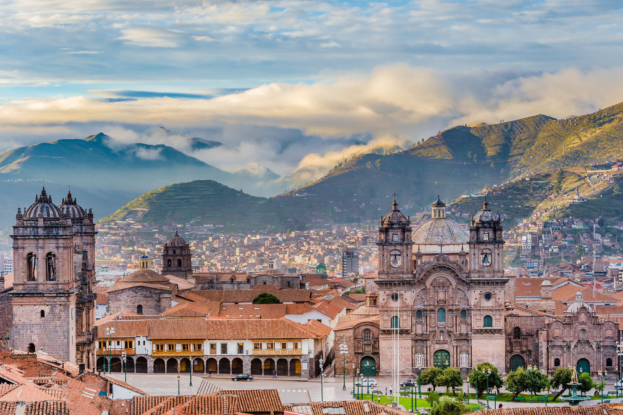 Elevated daytime view of Plaza de Armas in Cusco showing the Cusco Cathedral and Iglesia de la Compañía de Jesús flanking the main square, with colonial arcaded buildings, terracotta rooftops, and cloud-draped Andean mountains stretching into the distance, Peru.