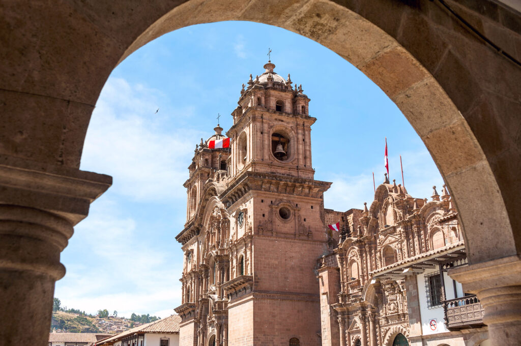 The ornate Andean Baroque facade and bell tower of the Iglesia de la Compañía de Jesús on Cusco's Plaza de Armas, Peru, framed through a colonial stone arch of the surrounding portal arcade, with Peruvian flags flying from the tower under a clear blue Andean sky.